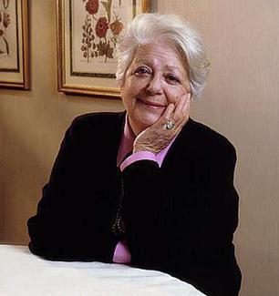 Elderly woman with white hair sits at a table, resting her chin on her hand and smiling, wearing a black cardigan over a pink top.