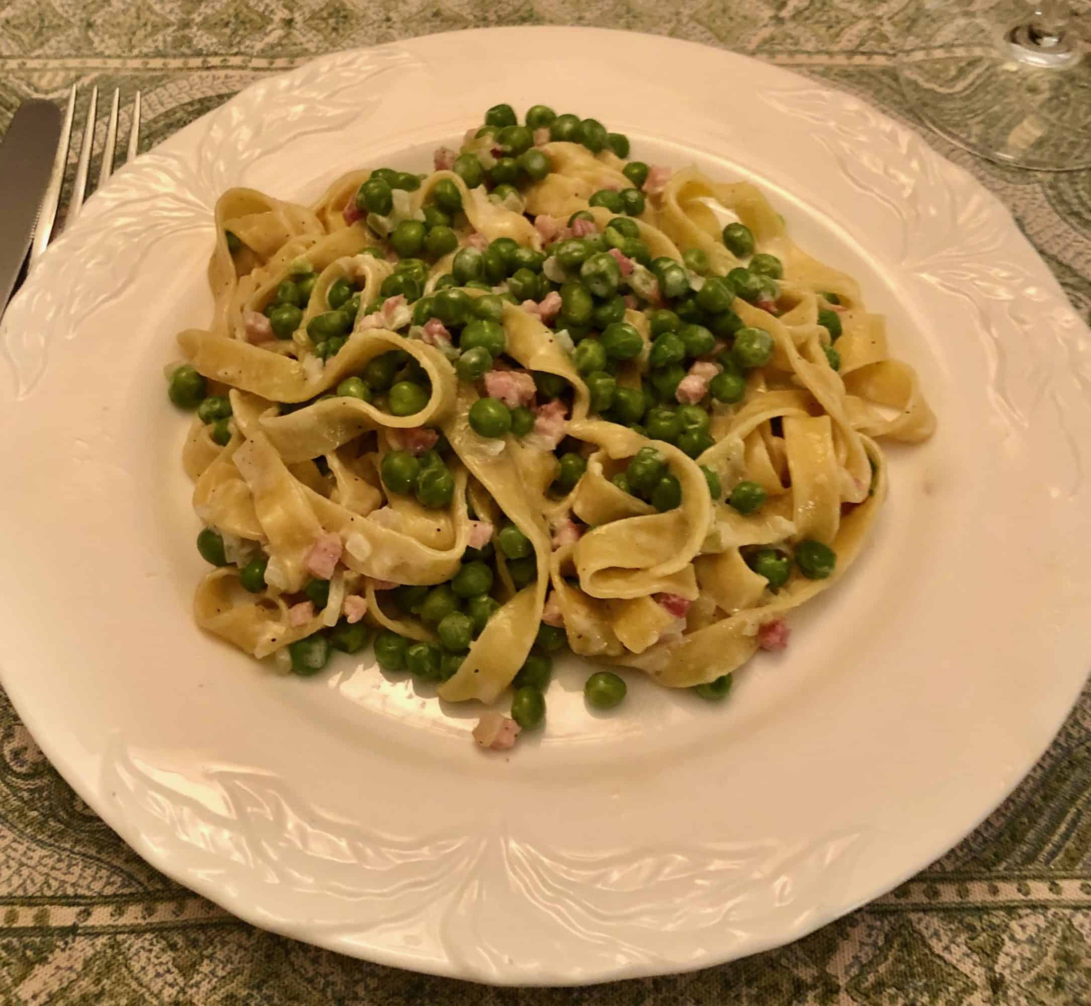 Plate of tagliatelle pasta with green peas and diced ham in a creamy sauce on a white decorative plate.