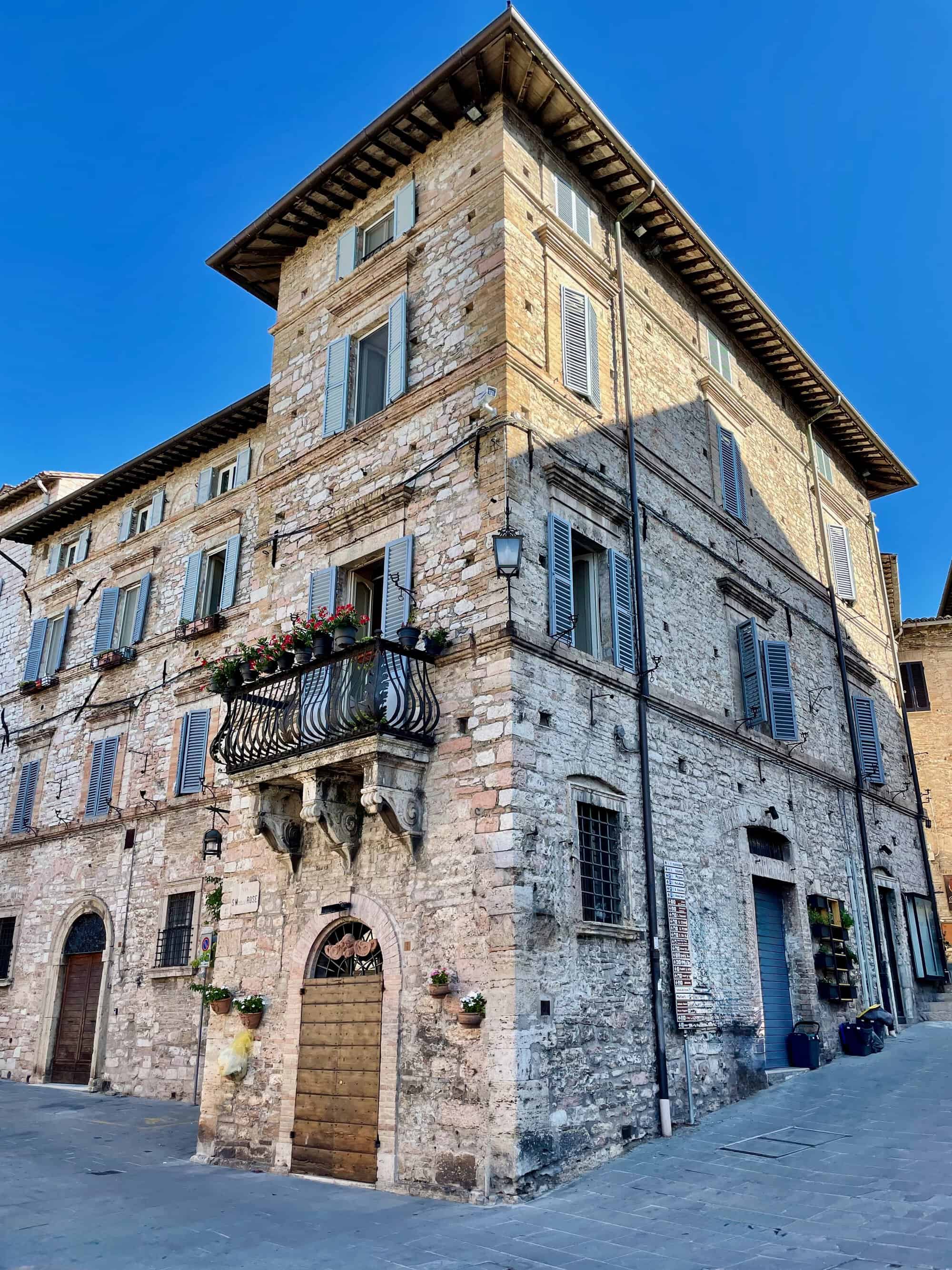 Corner stone building with blue shutters and a flower-filled balcony on a sunny street corner in a historic town.