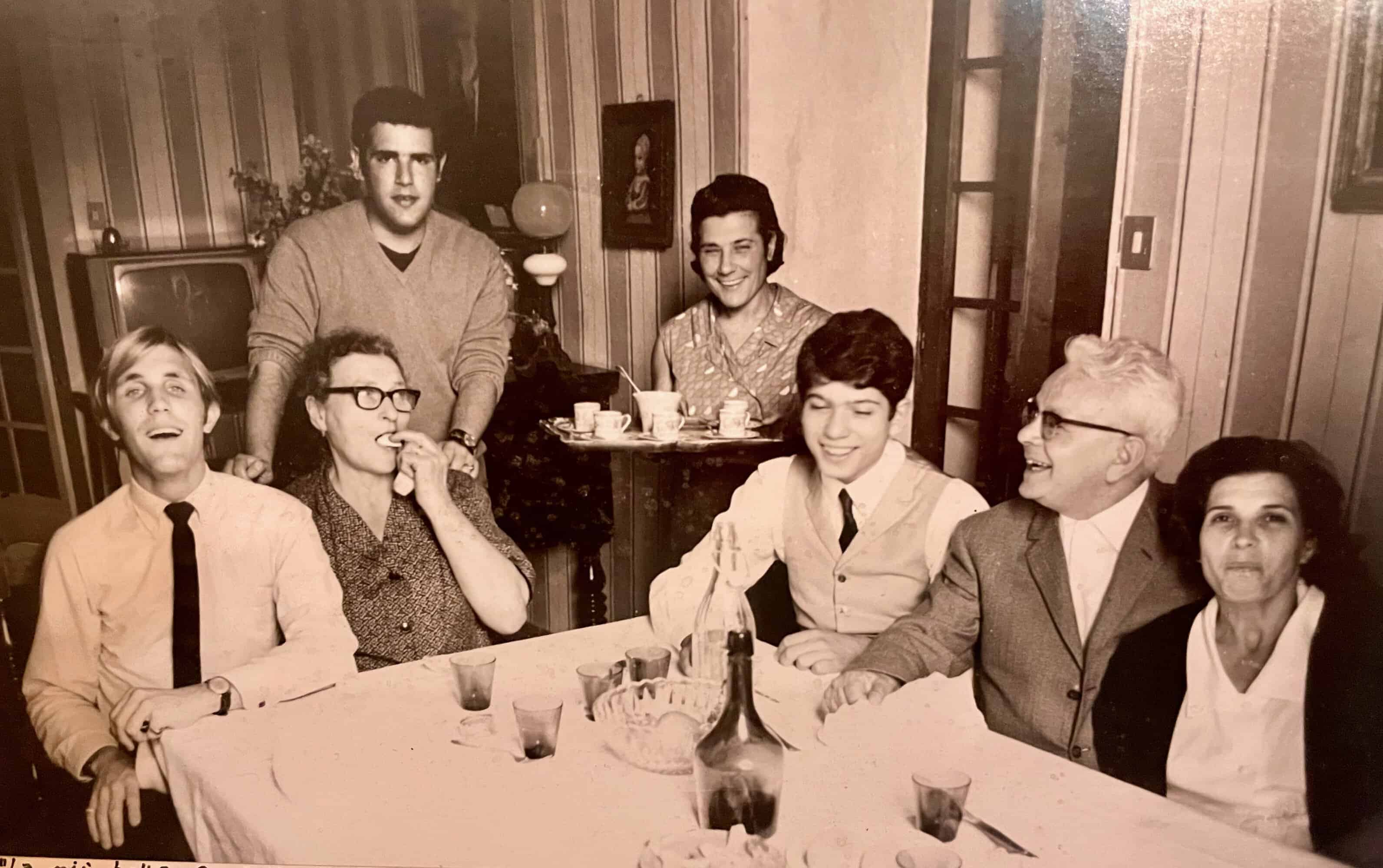 Six adults around a dining table in a mid‑century living room, smiling for a casual group photo.