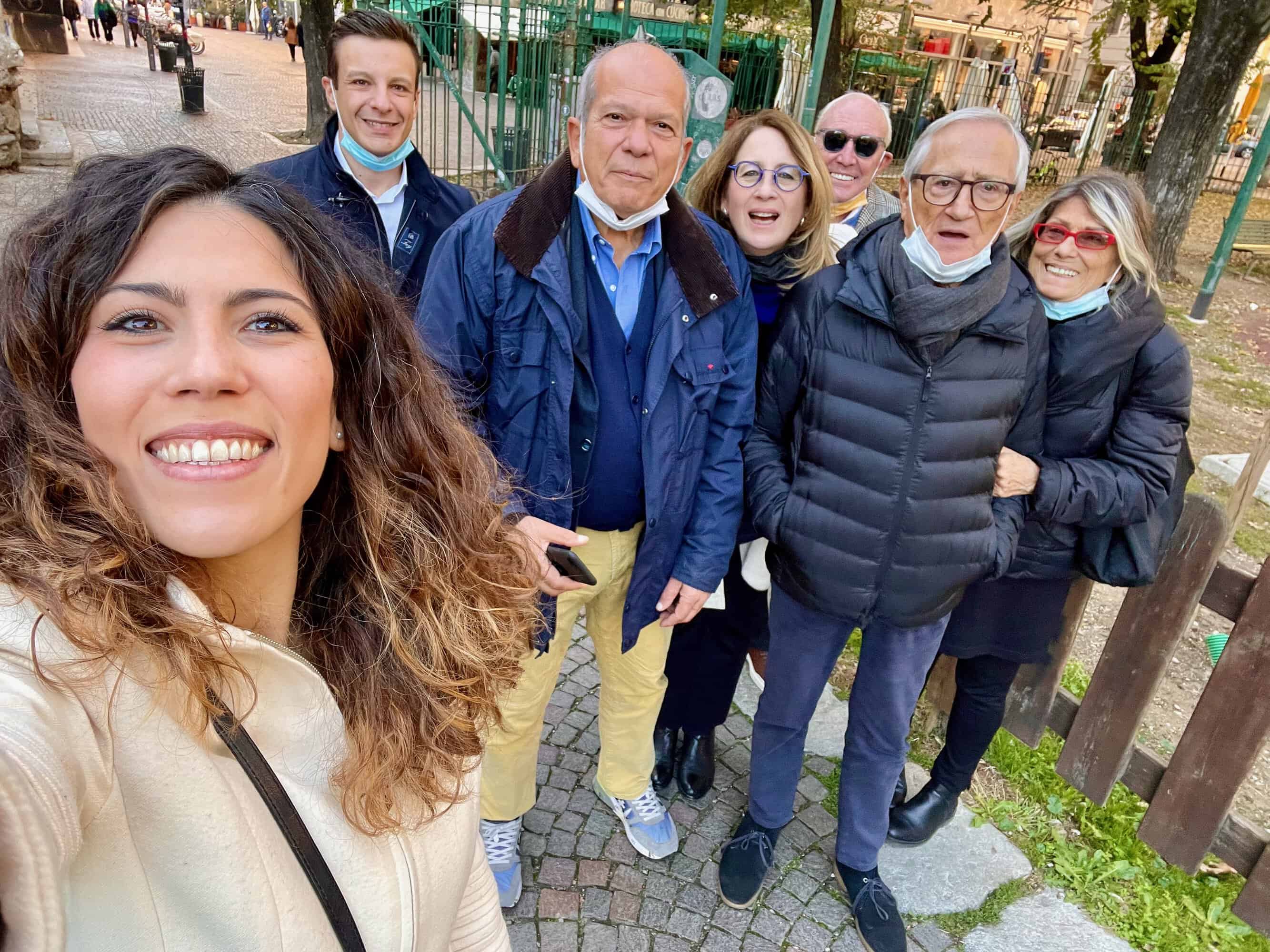 Woman at the left takes a selfie with a group of six people standing behind her on a cobblestone street in a park, everyone smiling and some wearing masks