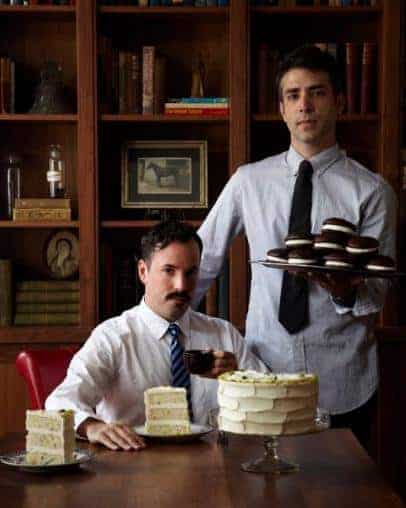 Two men in dress shirts and ties in a wood-paneled room, one seated with a cup and slices of cake on plates, the other standing and carrying a tray of stacked desserts.