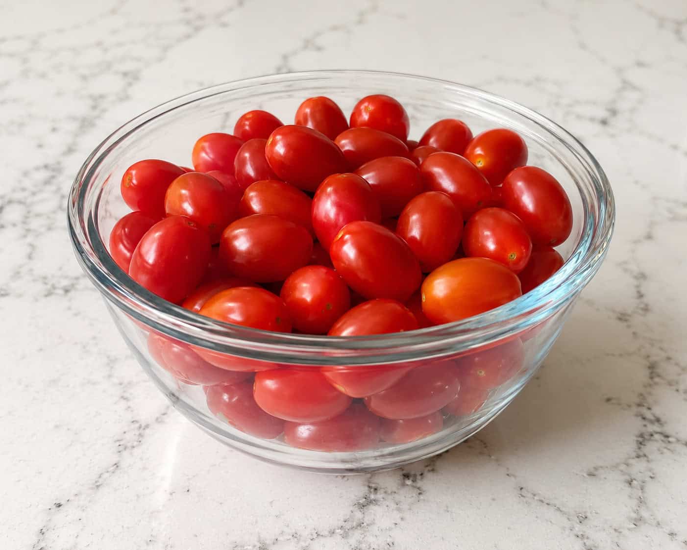 Glass bowl filled with red cherry tomatoes on a white marble countertop.