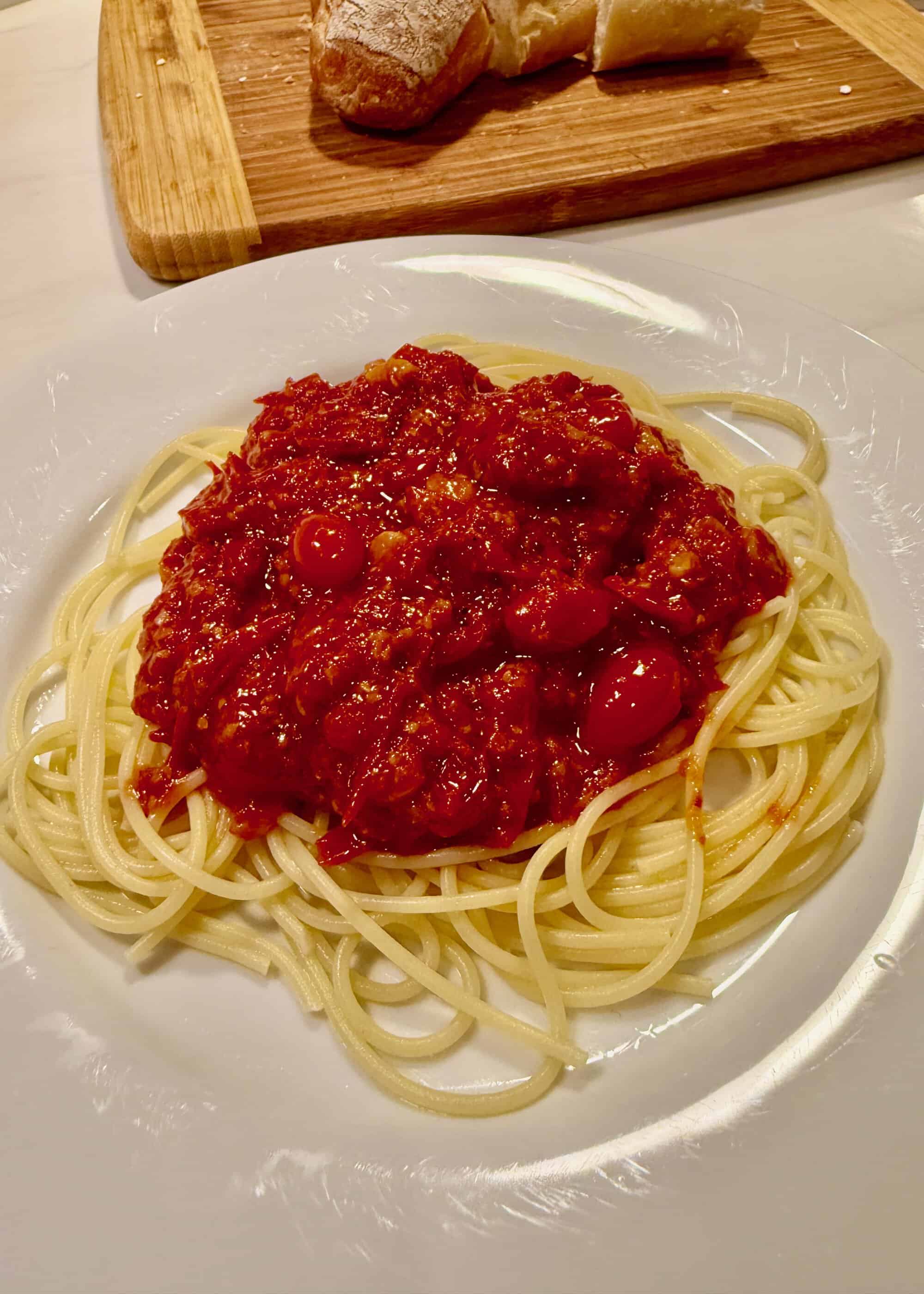 Plate of spaghetti with chunky red tomato sauce and olives, on a white dish with a wooden cutting board and sliced bread in the background.