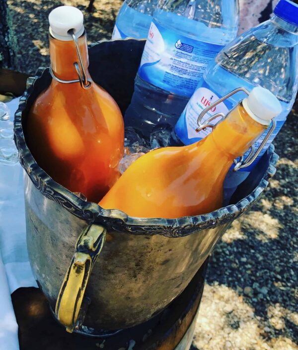 Close-up of orange glass bottles with white stoppers secured by a metal ring inside a metal bucket, next to blue plastic water bottles outdoors.