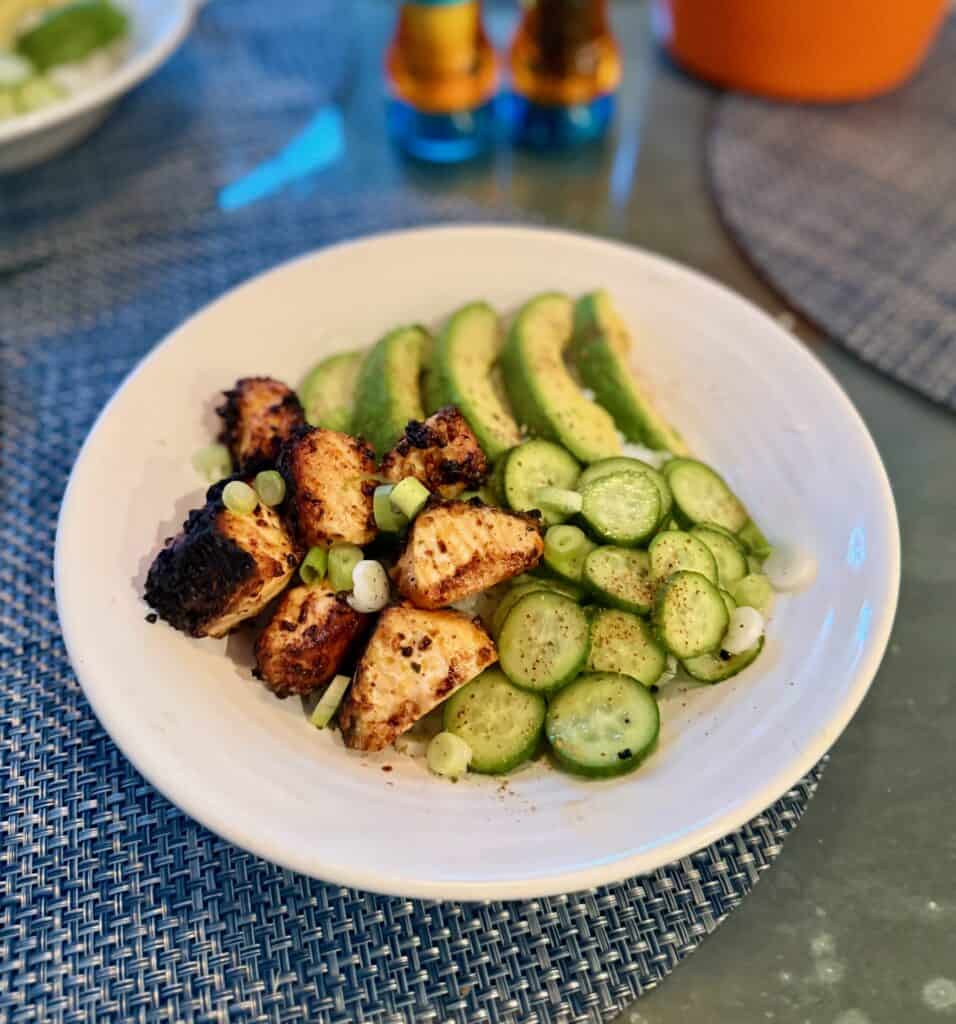 Plate with spicy salmon rice bowls and sliced cucumbers, garnished with green onions, on a white plate and blue table mat in a bright setting.