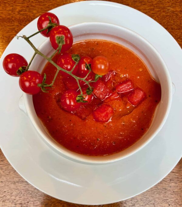 White bowl of bright red tomato soup with a vine of cherry tomatoes resting on the edge.