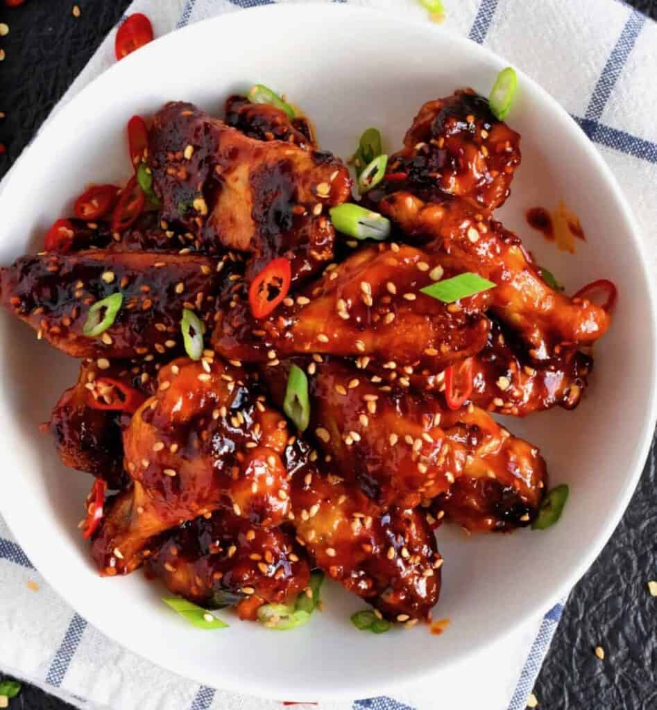 Sticky glazed chicken wings topped with sesame seeds and scallions in a white bowl.