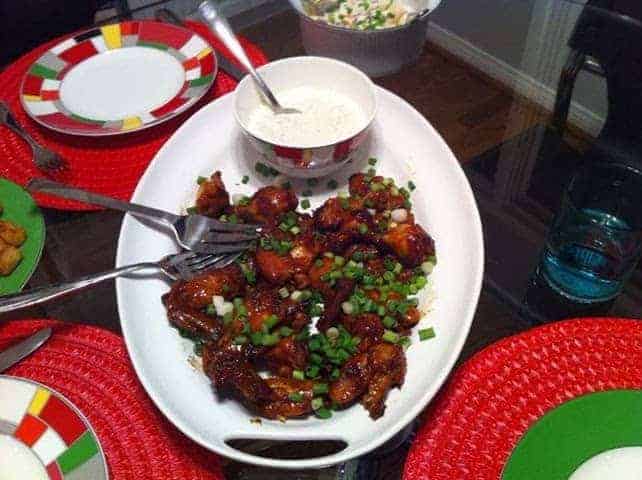Bowl of saucy glazed chicken bites topped with chopped scallions on a dining table with colorful plates and sauces nearby.