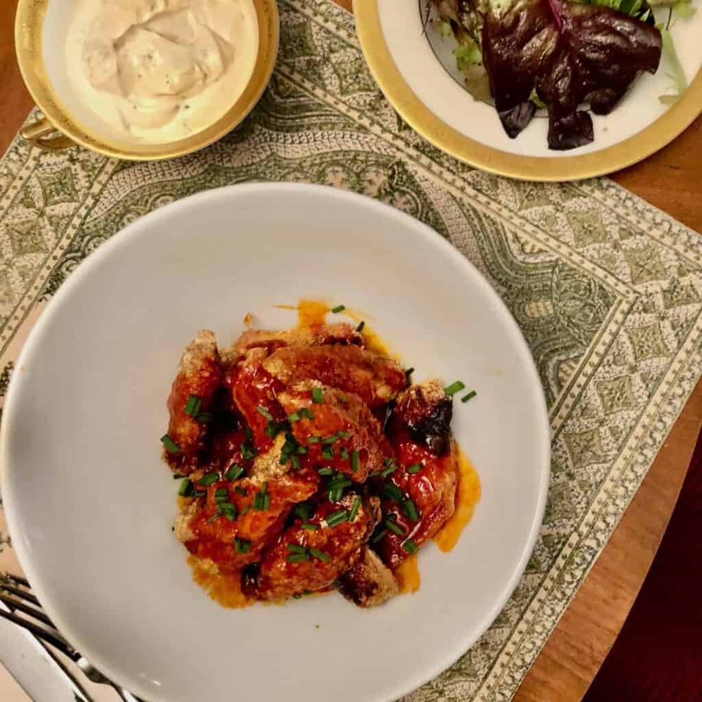 Plate of crispy fried chicken with tomato sauce, herbs, and chopped chives, served on a white dish with napkin pattern nearby.