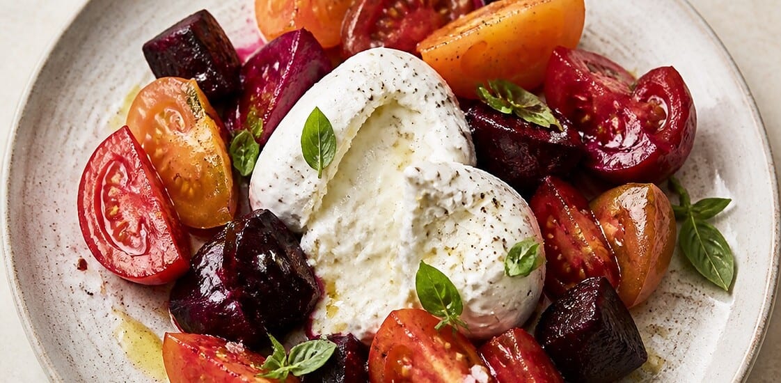 Burrata cheese on a plate surrounded by roasted beets and tomato wedges with fresh basil garnish.