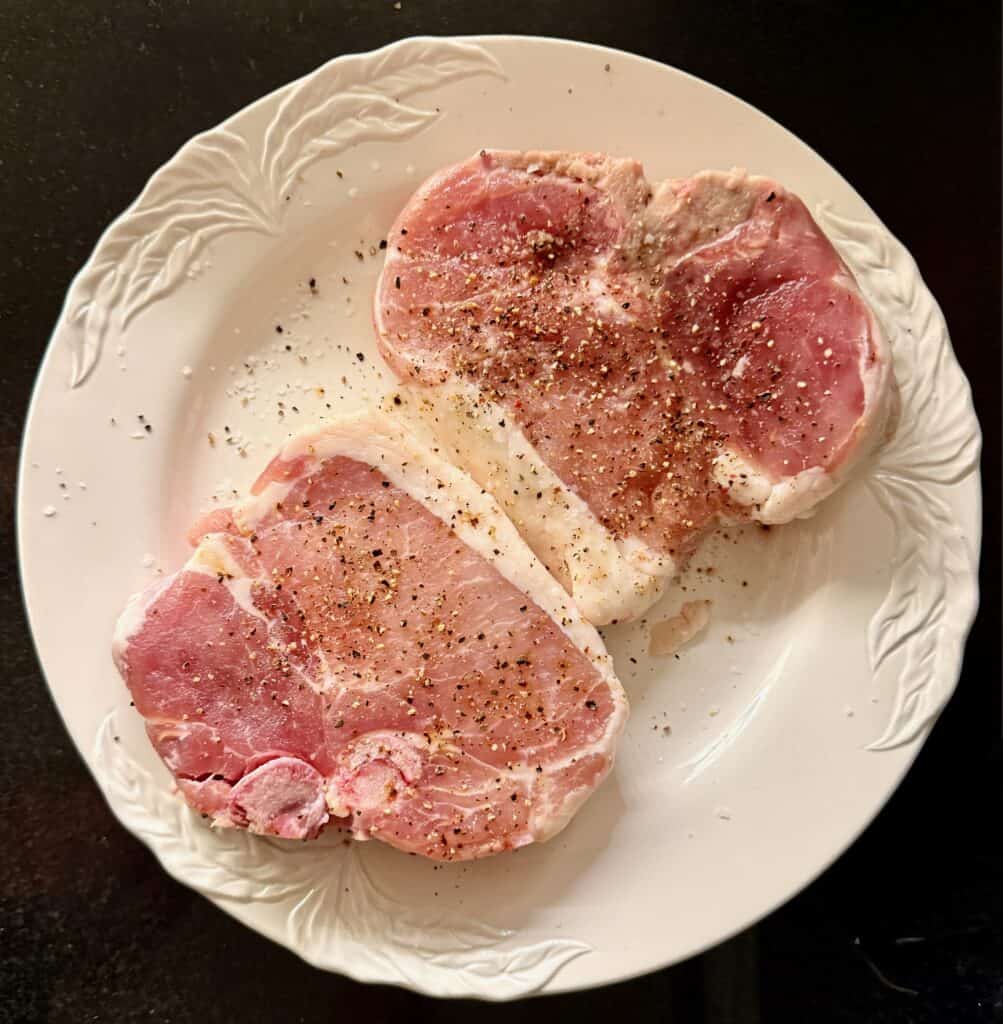 Two raw bone-in pork chops seasoned with salt and black pepper on a white plate.